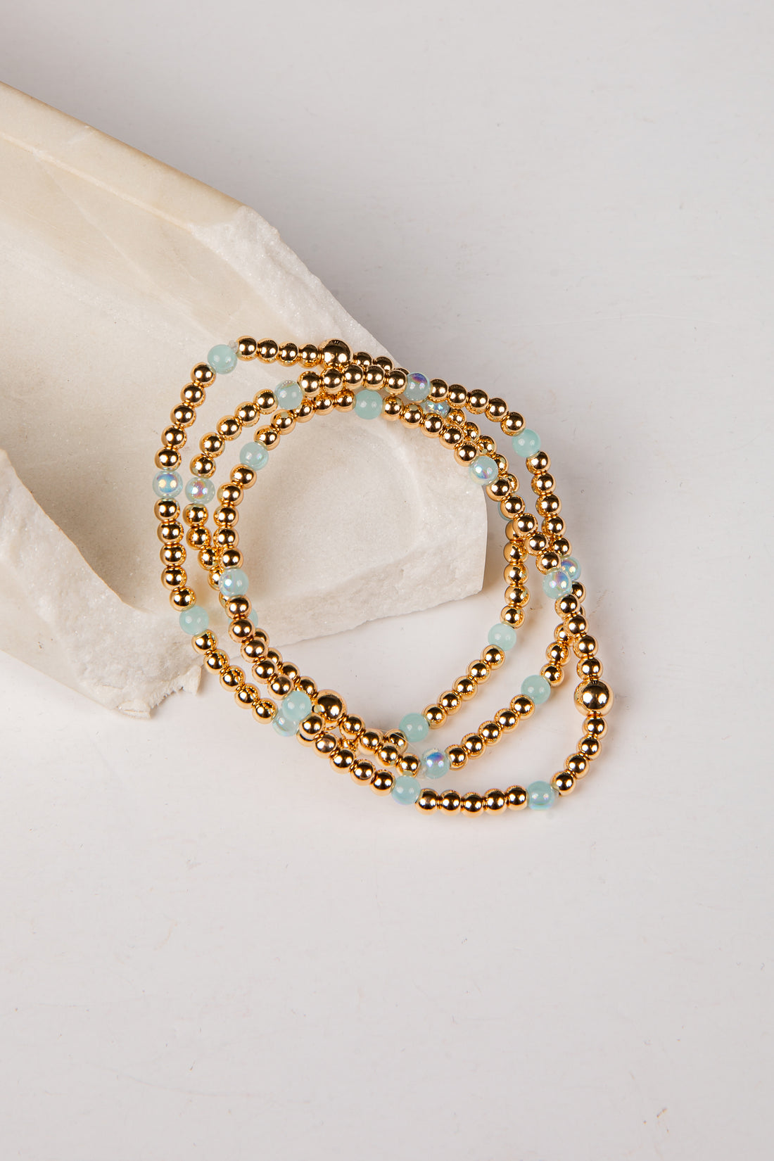 A multi-strand beaded necklace with metallic gold beads and light blue stone accents, styled on a white surface next to a cream-colored stone prop.