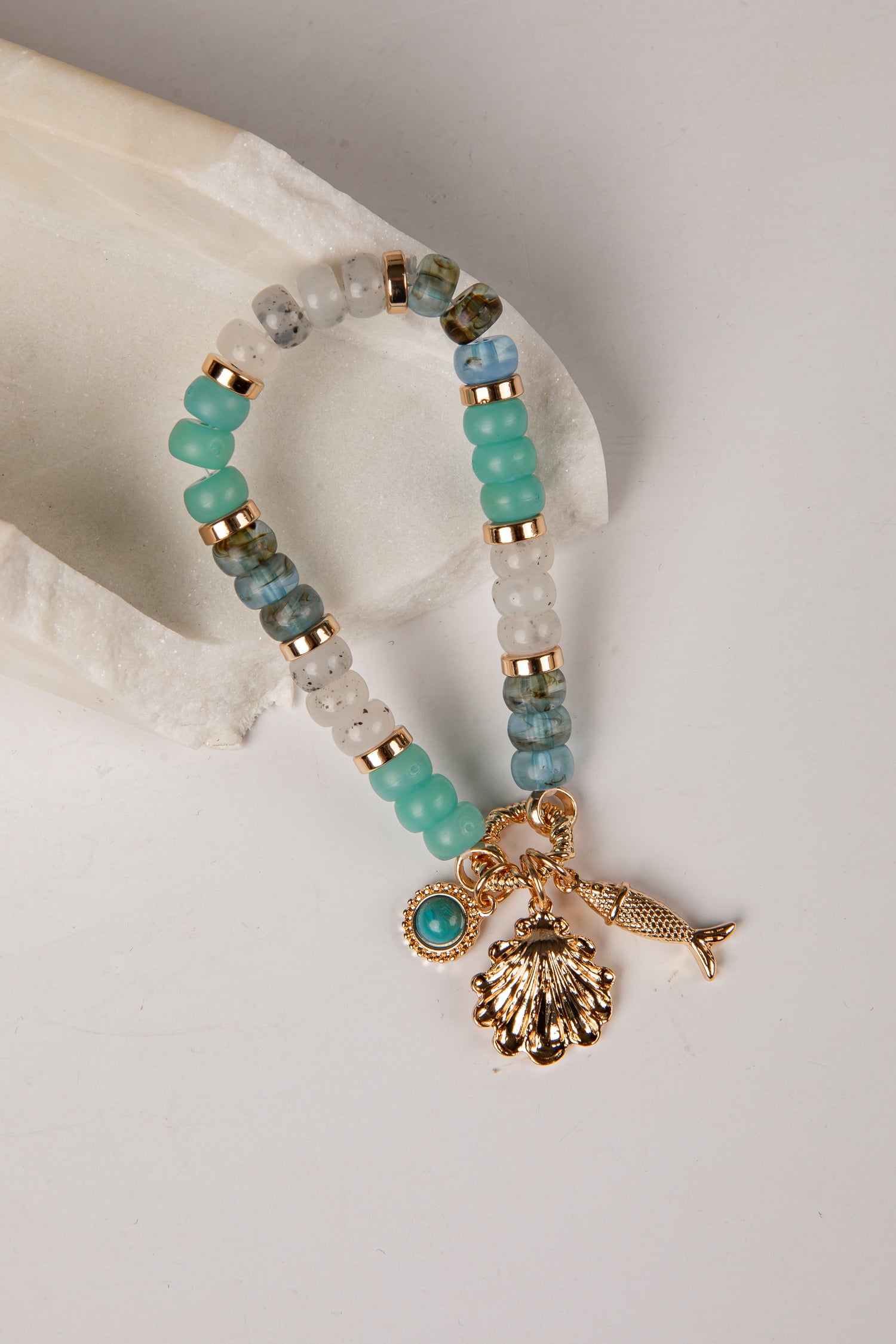 A beaded stretch bracelet with turquoise, white, and gray beads, gold spacer rings, and gold charms including a seashell, fish, and starfish, displayed on a white stone prop against a light gray background.