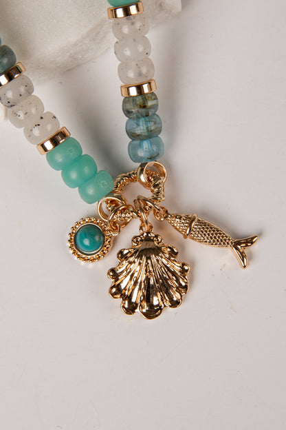 A close-up of a beaded jewelry charm strand with gold-tone metal charms, including a seashell, fish, and turquoise stone, alongside aqua and marbled beads on a white background.