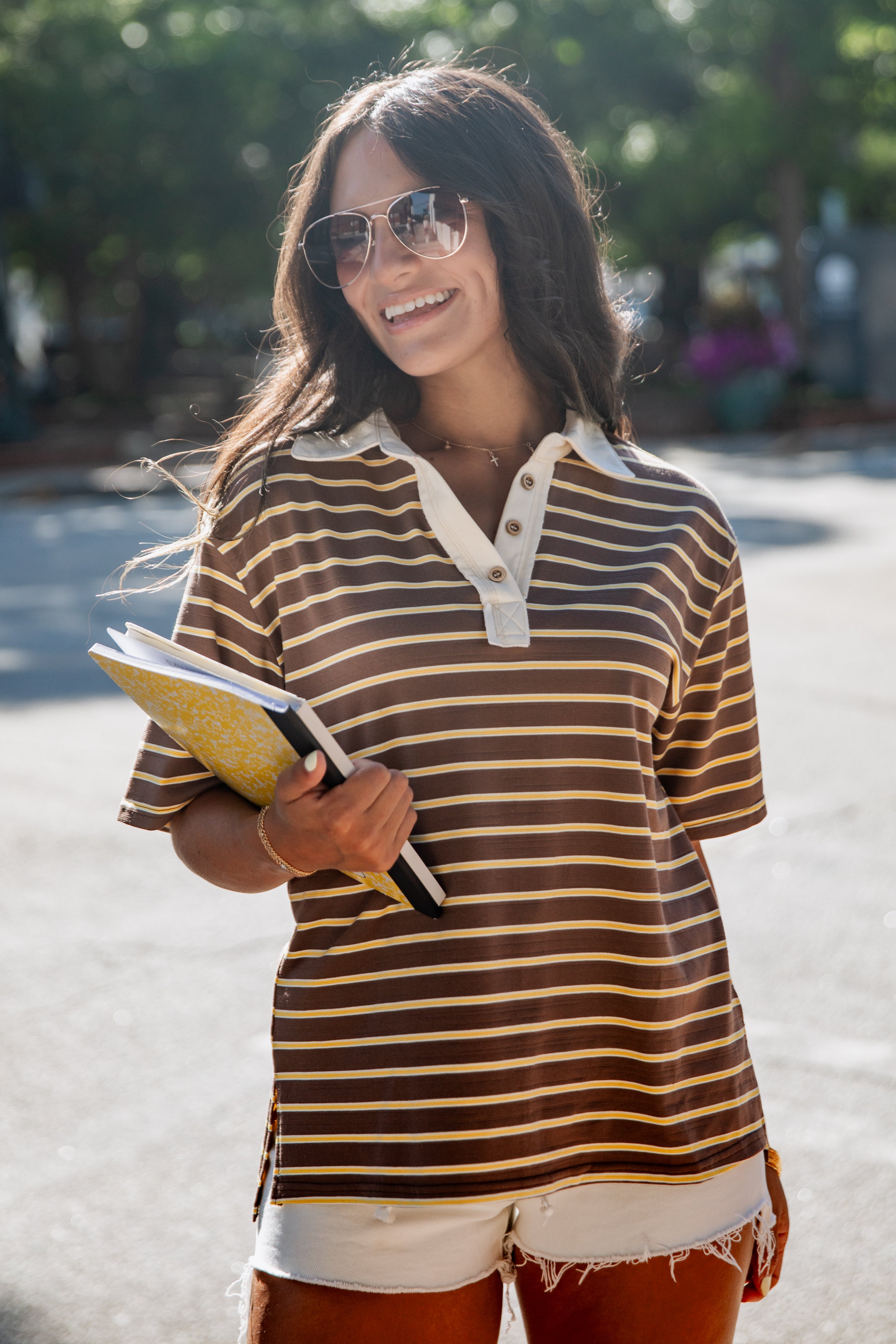 A woman wearing a brown and beige horizontally striped short-sleeve polo shirt with a white collar, holding a notebook in her hand, set against a blurred outdoor background.