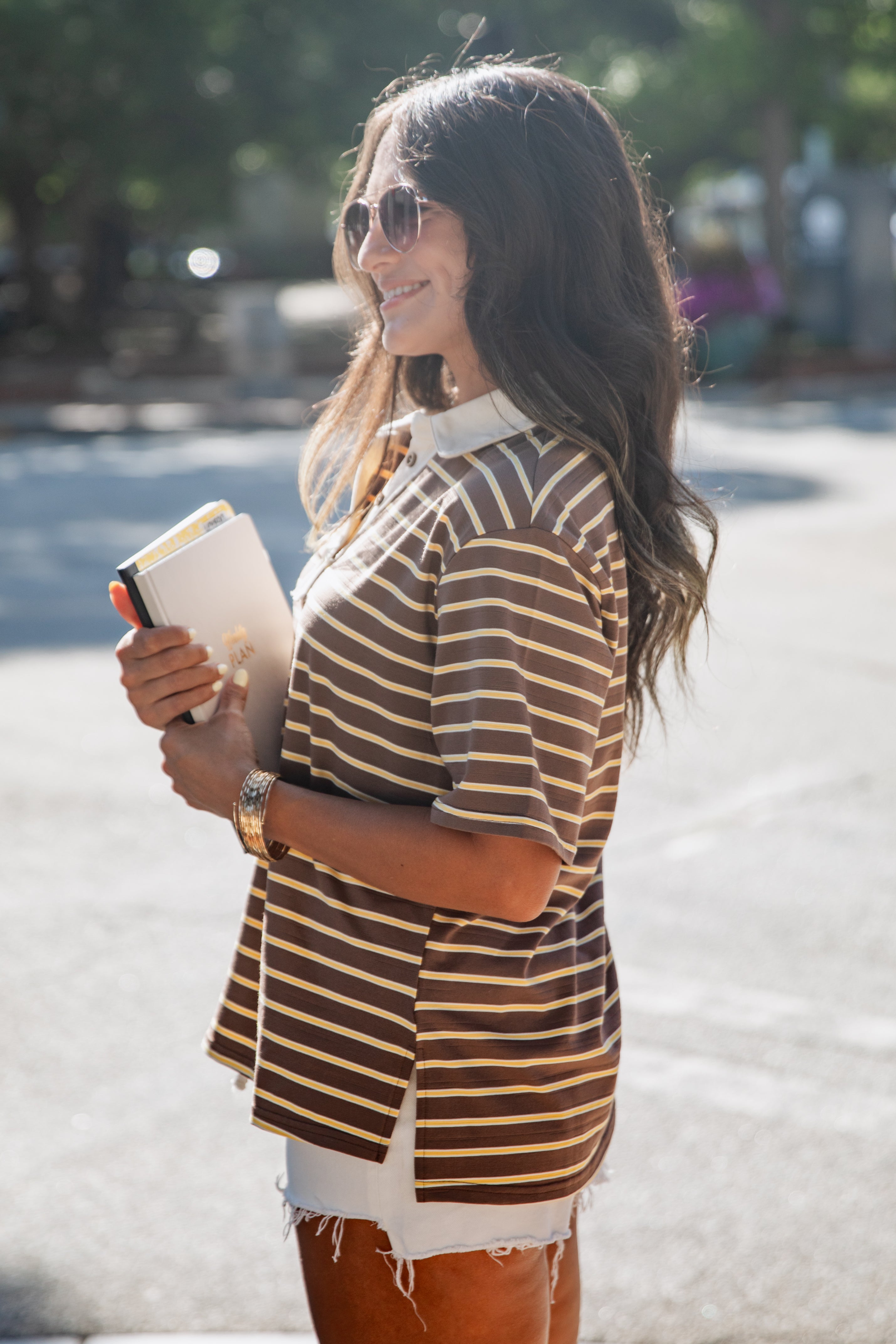 A woman wearing a brown and beige horizontally striped short-sleeve polo shirt, white shorts, and sunglasses, holding a notebook outdoors on a sunny day.