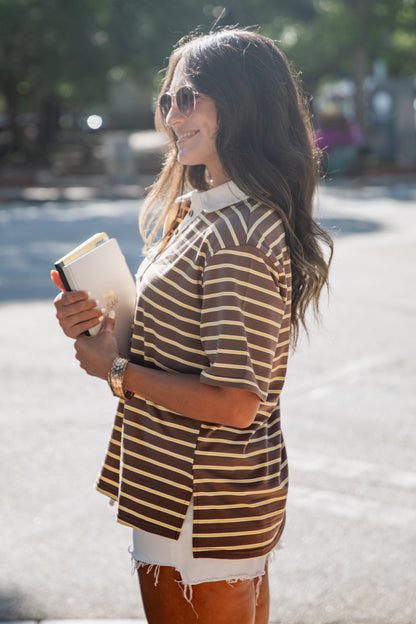 A woman wearing a brown and beige horizontally striped short-sleeve polo shirt, white shorts, and sunglasses, holding a notebook outdoors on a sunny day.