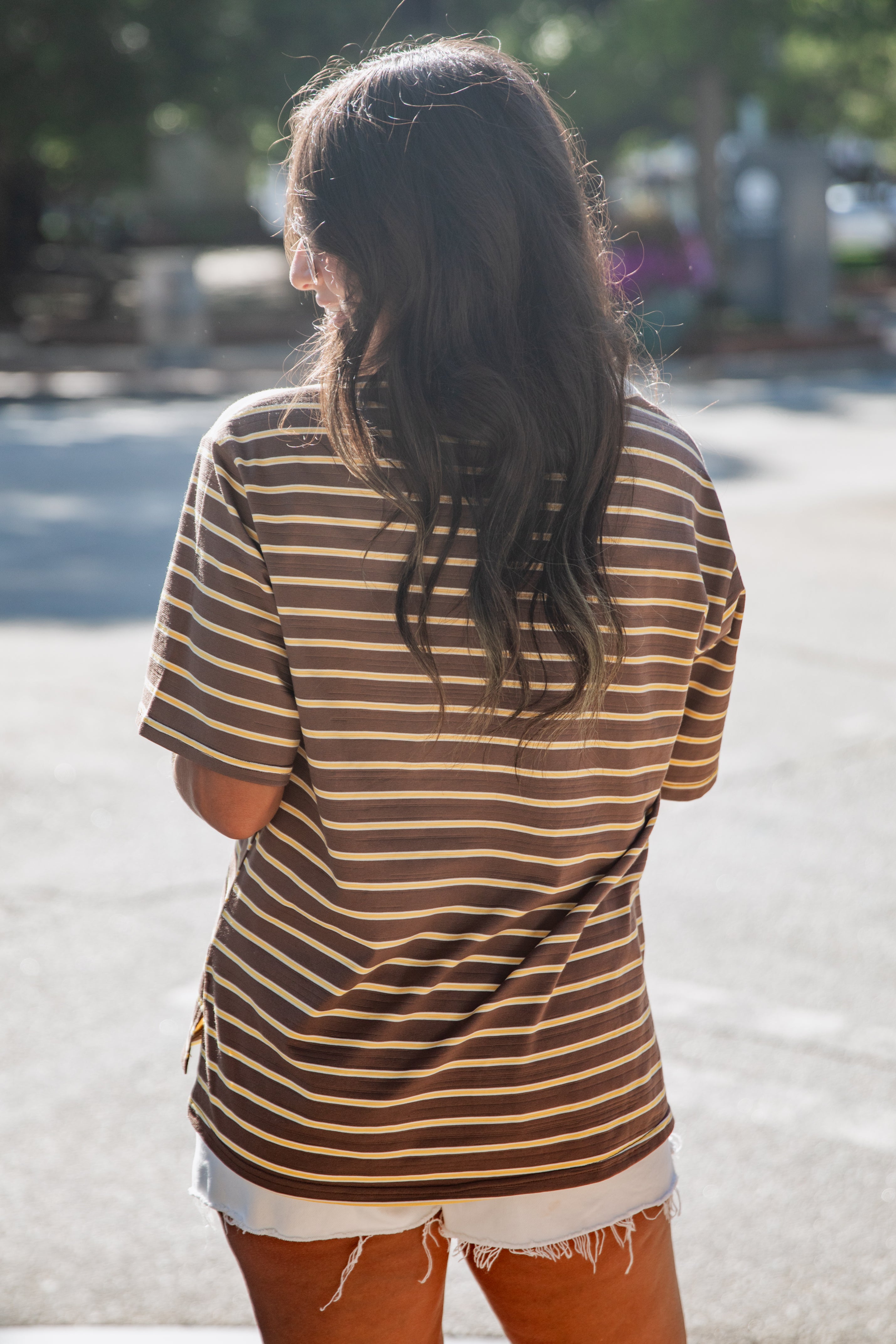 A woman viewed from behind wearing an oversized brown and tan striped t-shirt and white frayed denim shorts, standing outdoors on a sunny street.