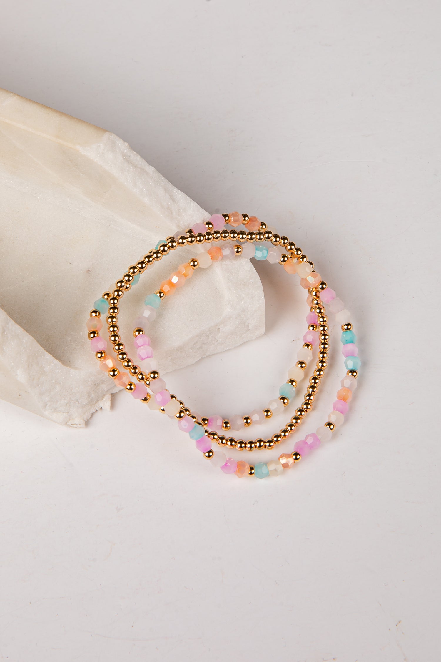 Three beaded stretch bracelets with gold-tone beads and pastel pink, peach, and mint blue beads, arranged on a white surface next to a white stone prop.