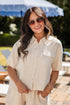 Woman wearing sunglasses and a beige outfit standing outdoors with greenery in the background