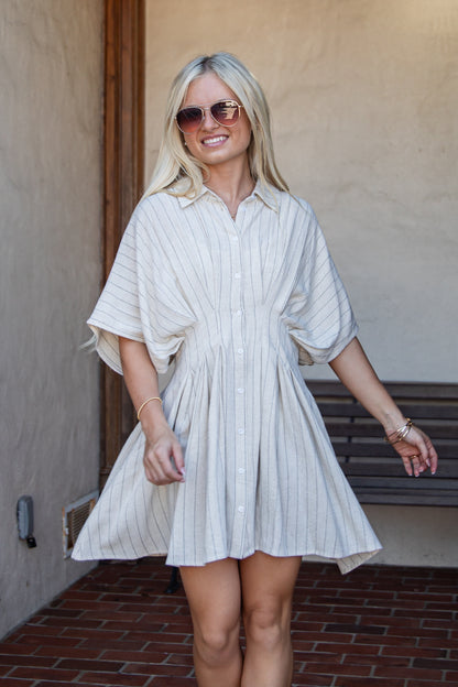 A woman wearing a cream and navy blue vertically striped button-down shirt dress, standing in front of a stucco wall and brick floor.