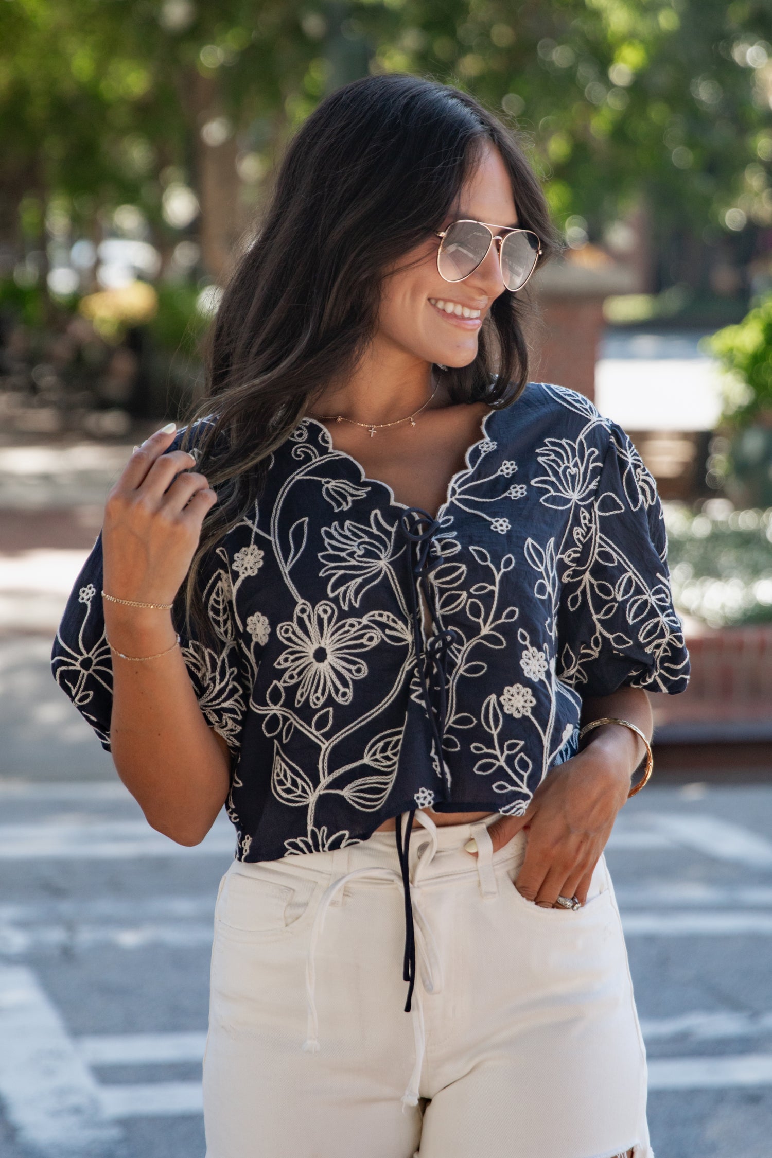 A woman wearing a navy blue short-sleeve blouse with a white floral pattern and cream drawstring shorts, standing outdoors on a sunny day.