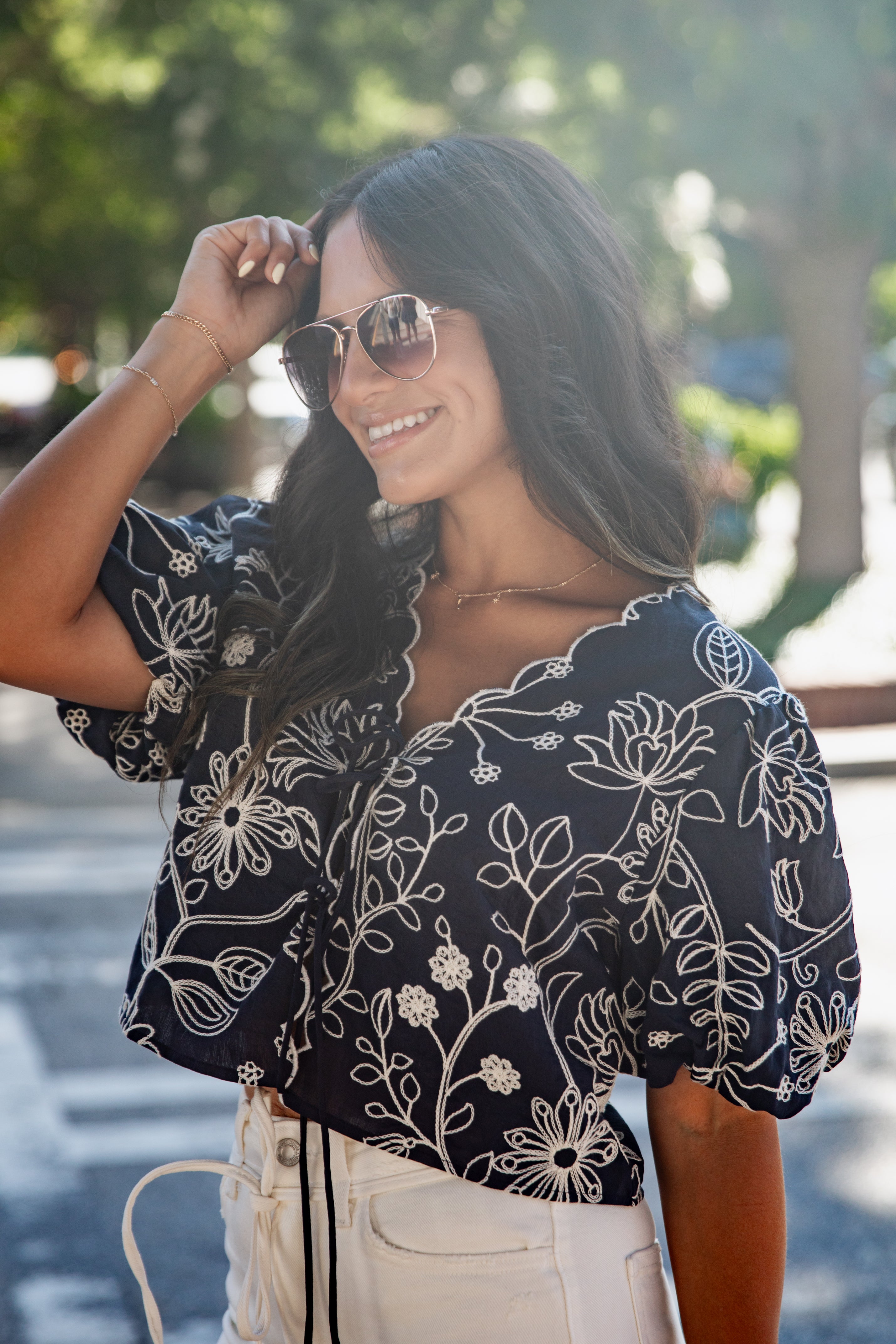 A woman wearing a navy blue short-sleeve blouse with a white floral print and puff sleeves, paired with cream-colored pants, photographed outdoors in a sunny, blurred street setting.