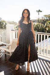 Woman in a black polka dot dress standing on a balcony with a scenic background.