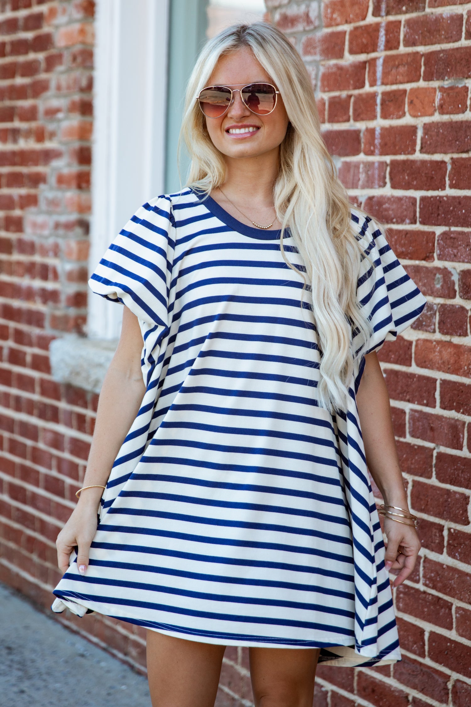 A woman with long blonde hair and sunglasses wearing a blue and white horizontally striped tunic dress, standing in front of a red brick wall.