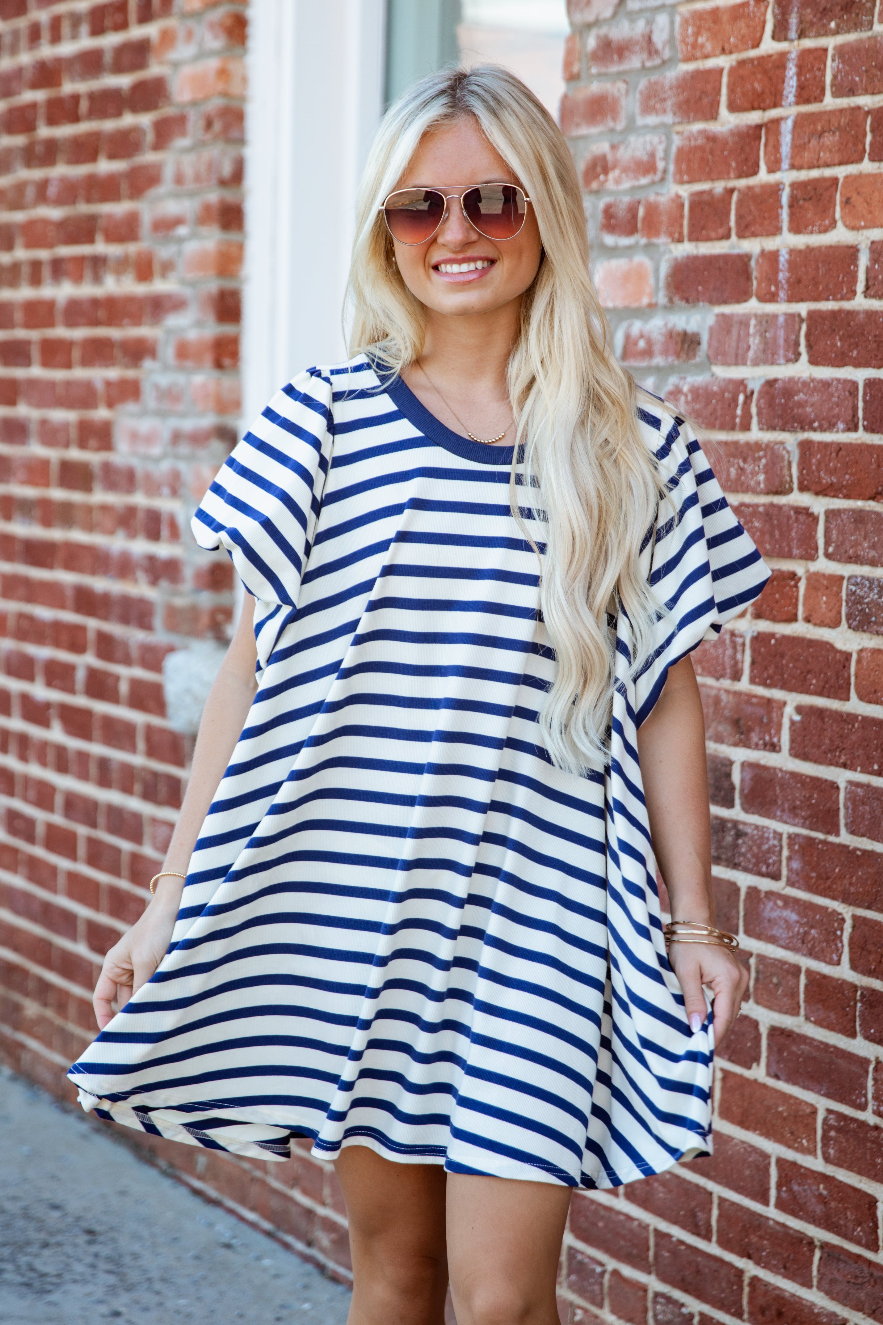 A woman with long blonde hair and sunglasses wearing a blue and white striped tunic dress, standing in front of a red brick wall.