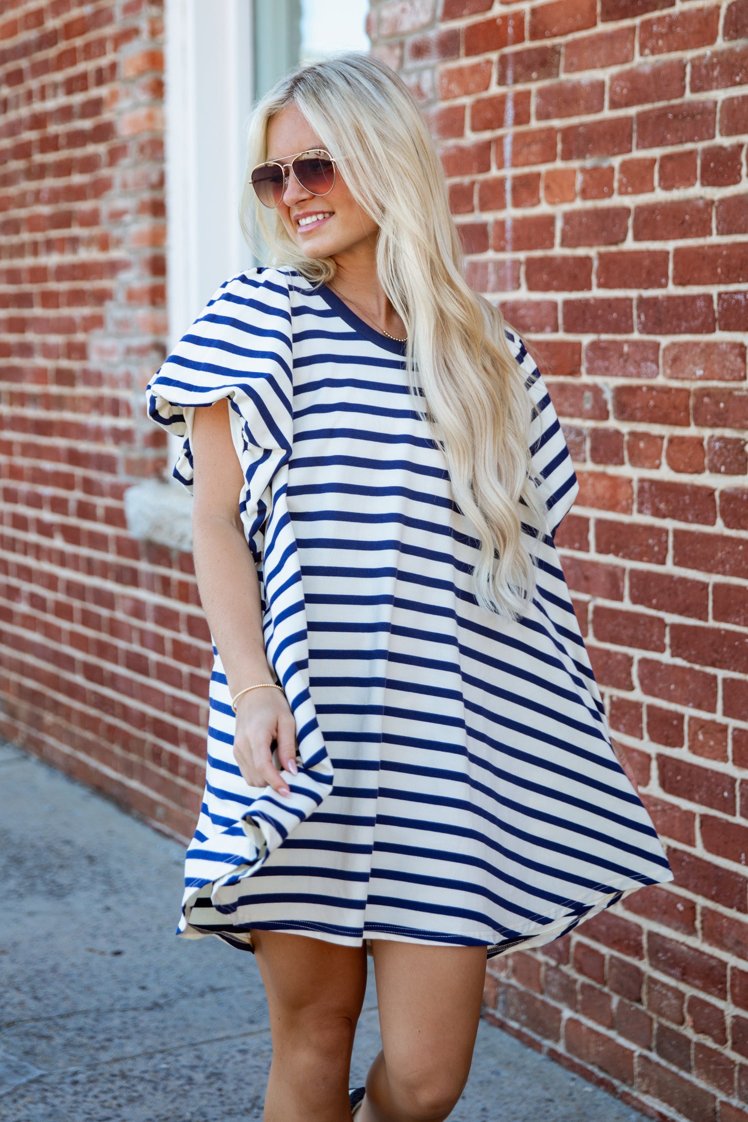 A woman with long blonde hair and sunglasses wearing a blue and white striped tunic dress, standing outdoors against a red brick wall.