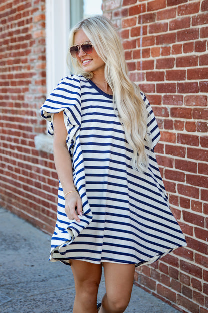 A woman with long blonde hair and sunglasses wearing a blue and white striped tunic dress, standing outdoors against a red brick wall.