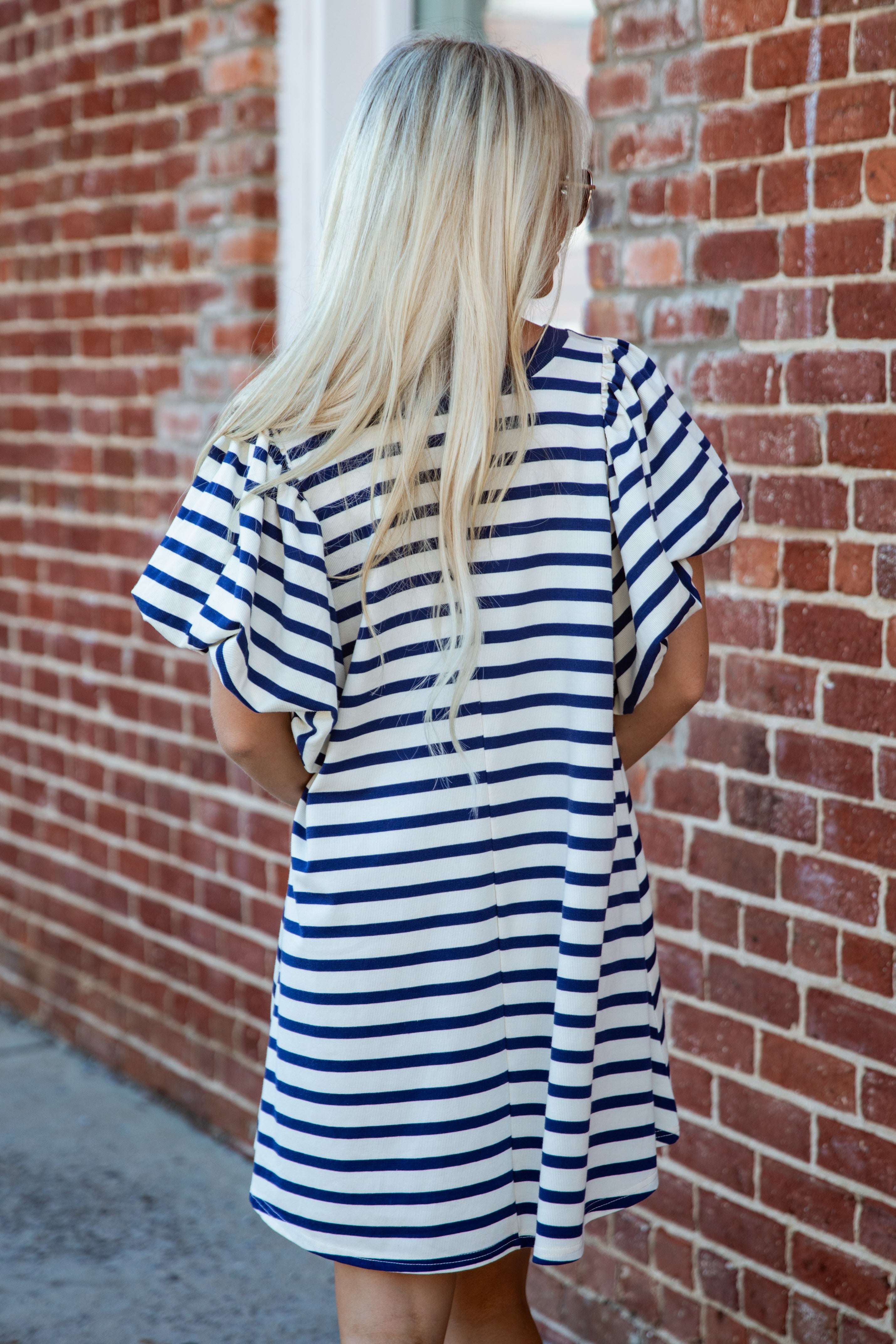 A woman with long blonde hair is shown from behind wearing a short-sleeve, navy and white horizontally striped tunic dress, standing in front of a red brick wall.