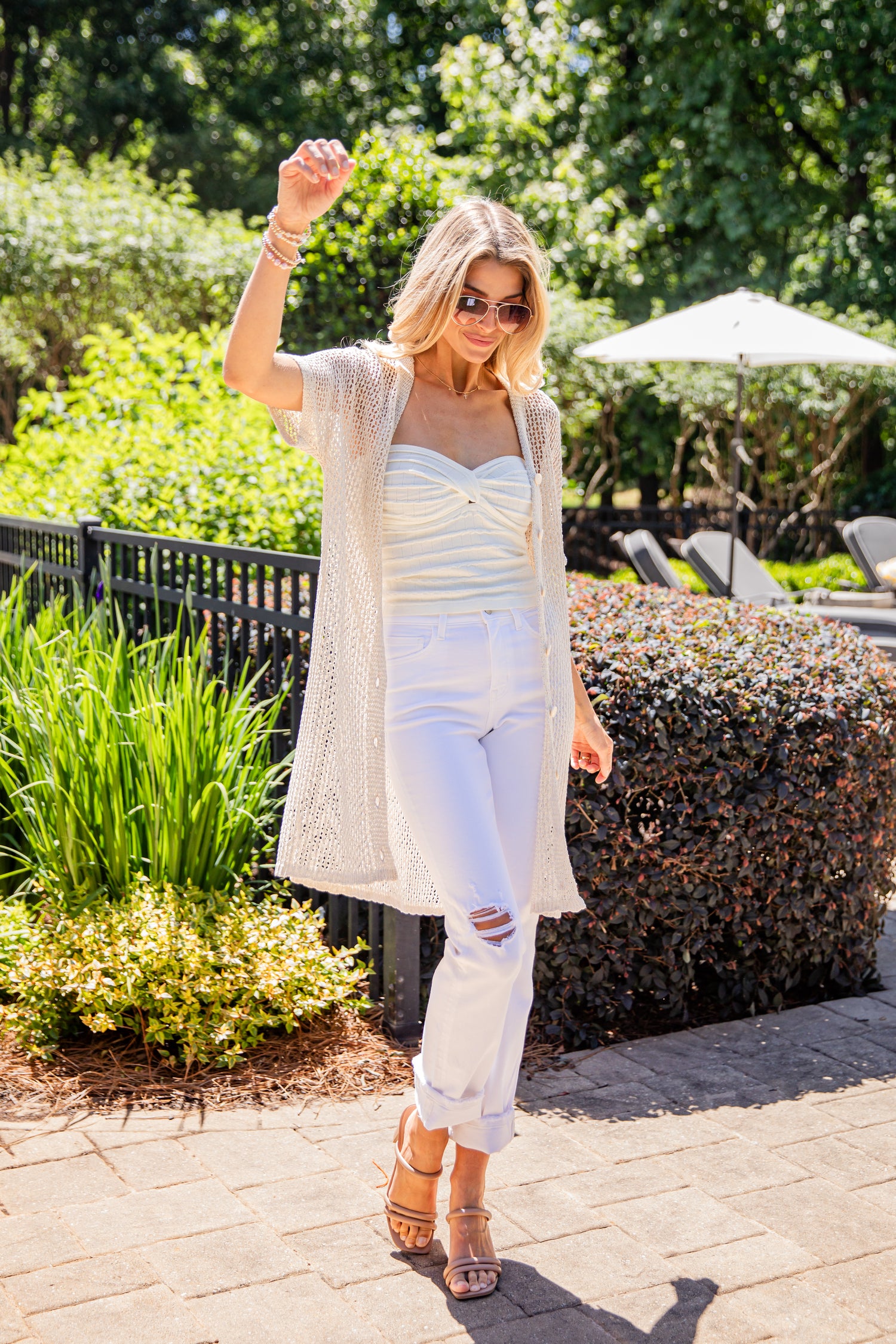 Woman in a white outfit with a beige cardigan standing outdoors with greenery in the background