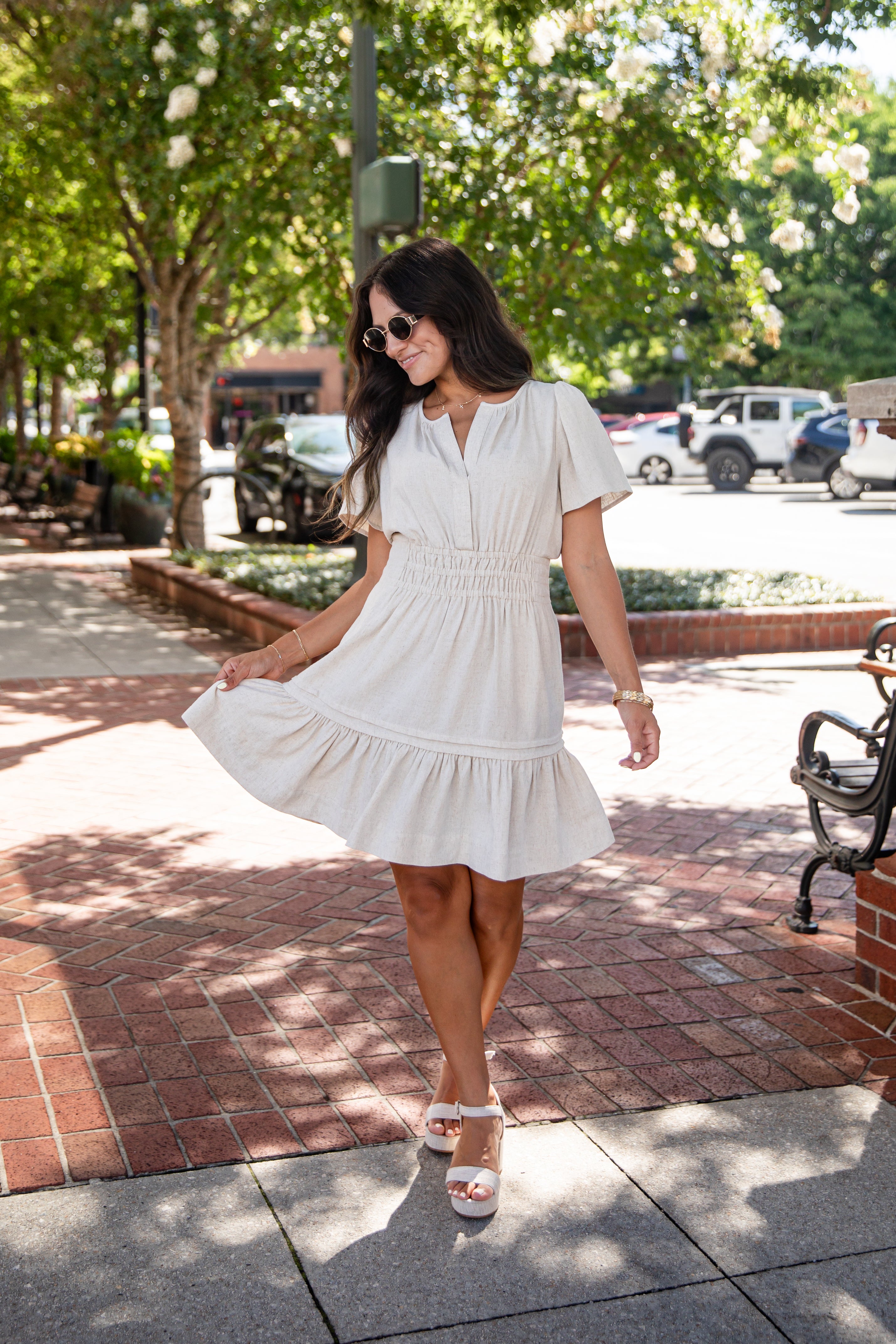 A woman wearing a short-sleeve white tiered dress and white platform sandals walking on a brick sidewalk in a sunny, tree-lined urban setting.