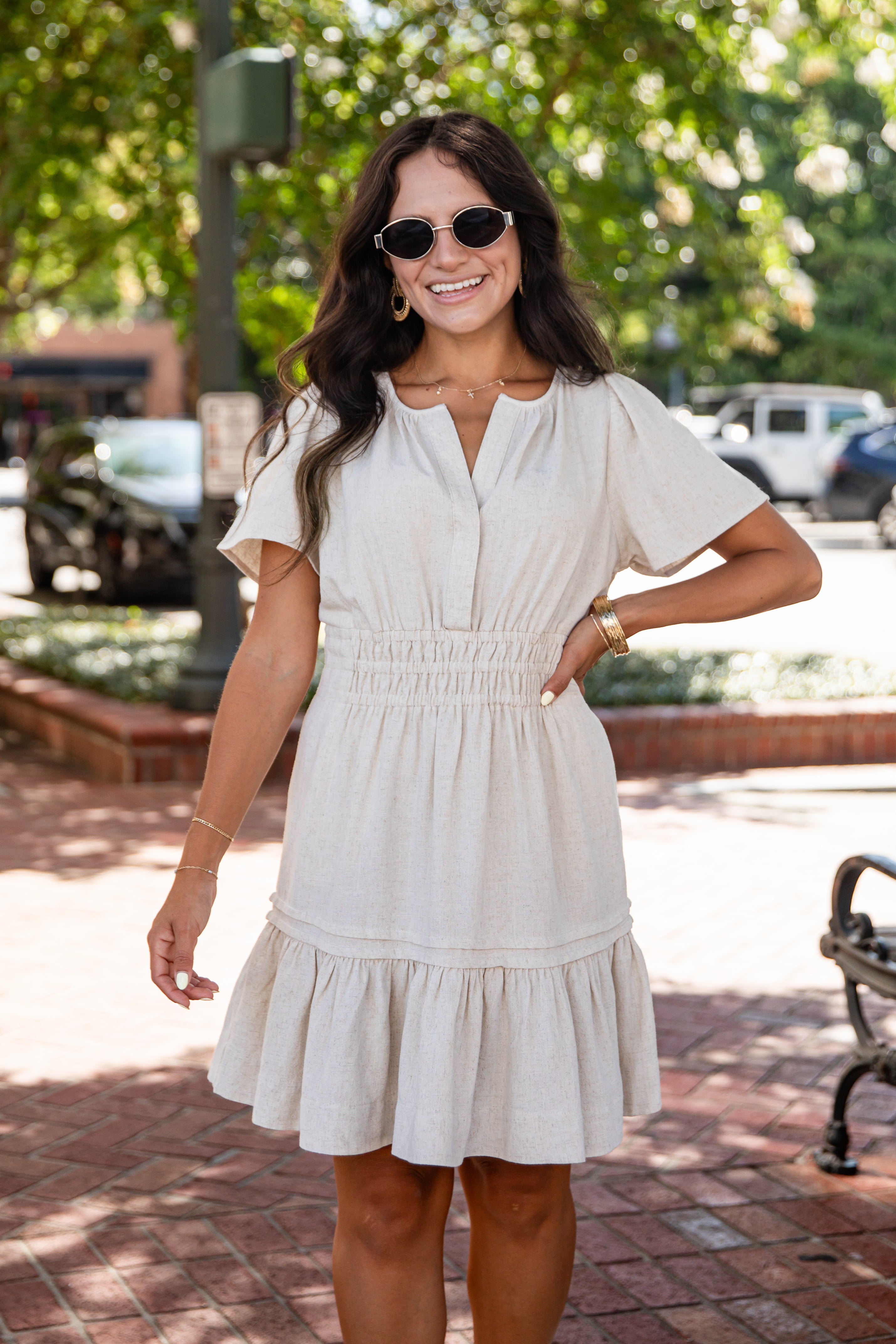 A woman wearing a short-sleeve cream linen dress with a smocked waist and tiered skirt, standing outdoors on a brick walkway with trees in the background.