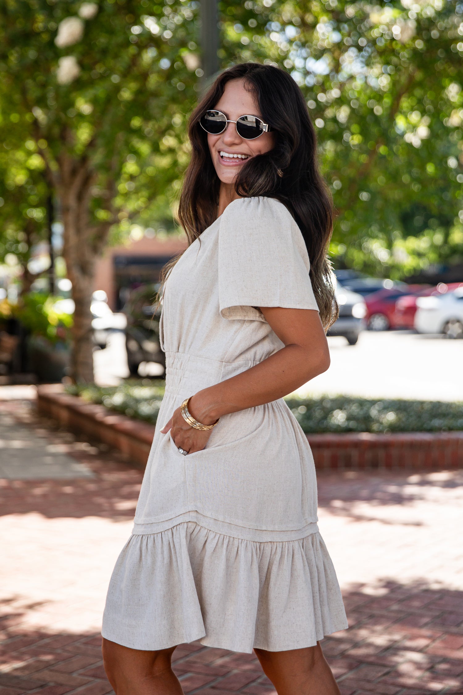 A woman wearing a light beige short-sleeve tiered dress and round sunglasses, posing outdoors on a brick walkway with trees and parked cars in the background.