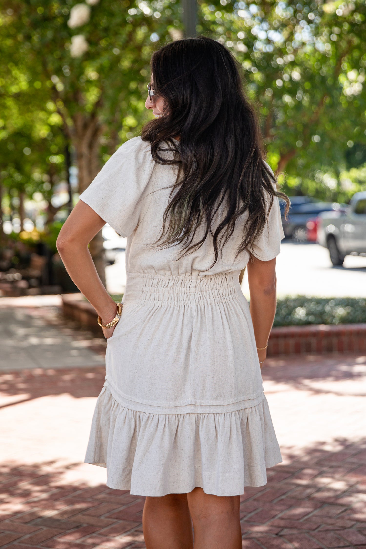 A woman viewed from behind wearing a short-sleeve cream-colored dress with a smocked waist and tiered skirt, standing outdoors on a brick walkway with trees and parked cars in the background.