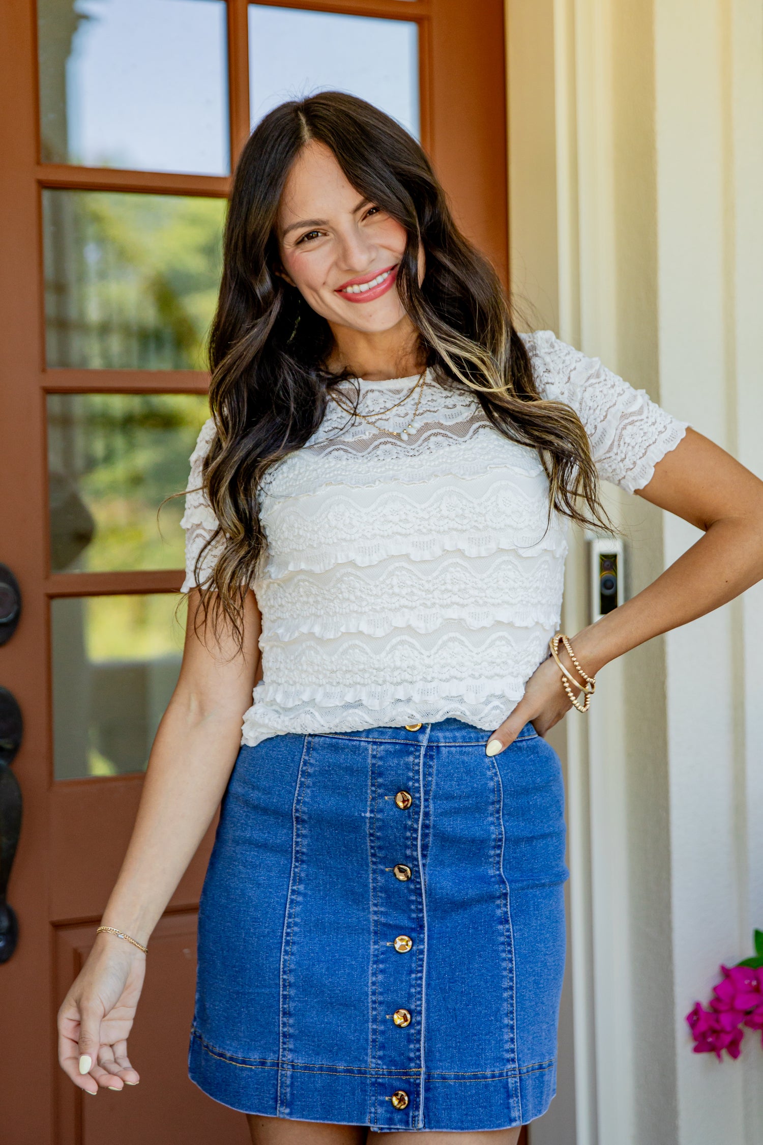 A woman wearing a white lace short-sleeve top and a blue denim button-front mini skirt, standing in front of a brown door and beige wall.