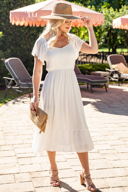 Woman wearing a white midi dress with flutter sleeves, a smocked bodice, and a tiered skirt, styled with a woven clutch, straw hat, and heeled sandals. The airy silhouette and timeless design create an effortlessly elegant look for summer vacations, bridal events, and special occasions.