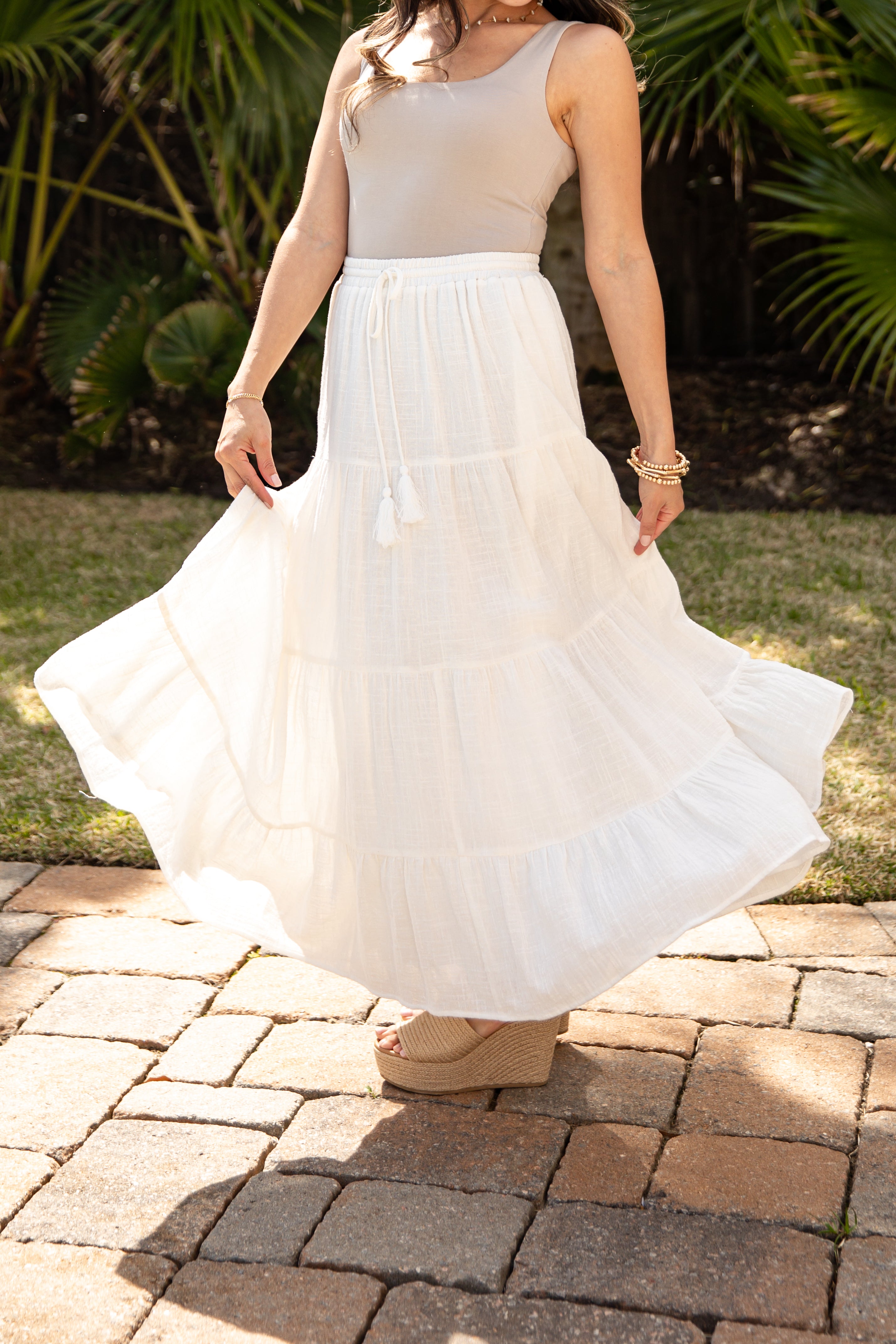 Woman wearing a beige top and white skirt standing on a paved path with greenery in the background