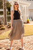 Woman in black top and beige skirt standing on a brick path with a house and trees in the background