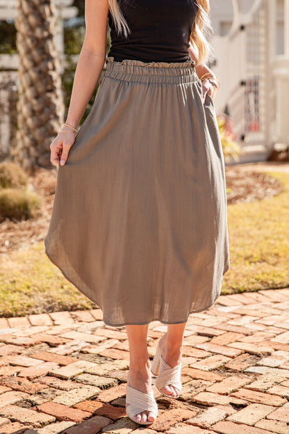 Person wearing a beige skirt and black top on a brick path with grass and trees in the background