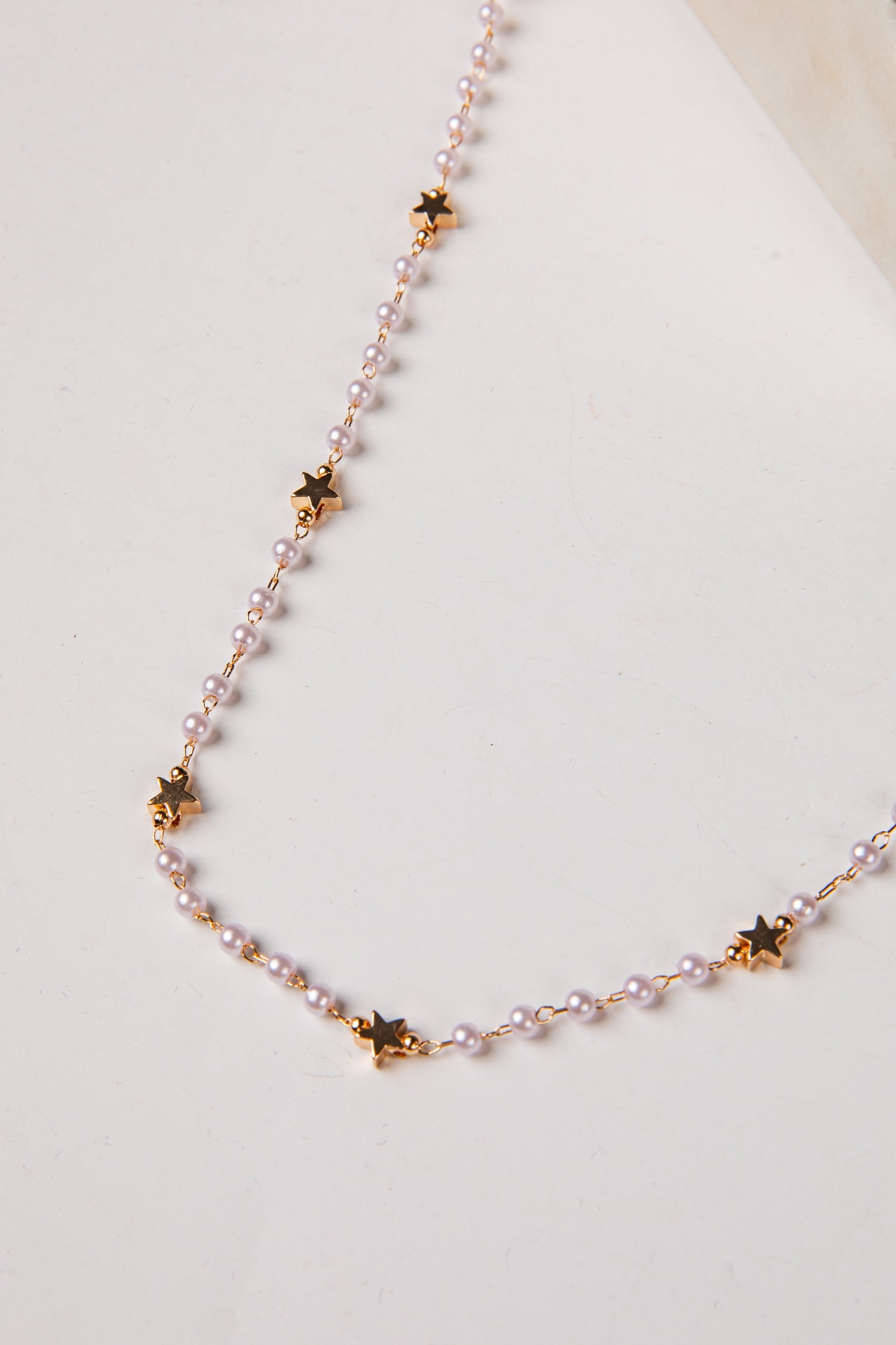 A delicate necklace featuring small light pink pearls and gold star-shaped charms, displayed on a clean off-white surface.