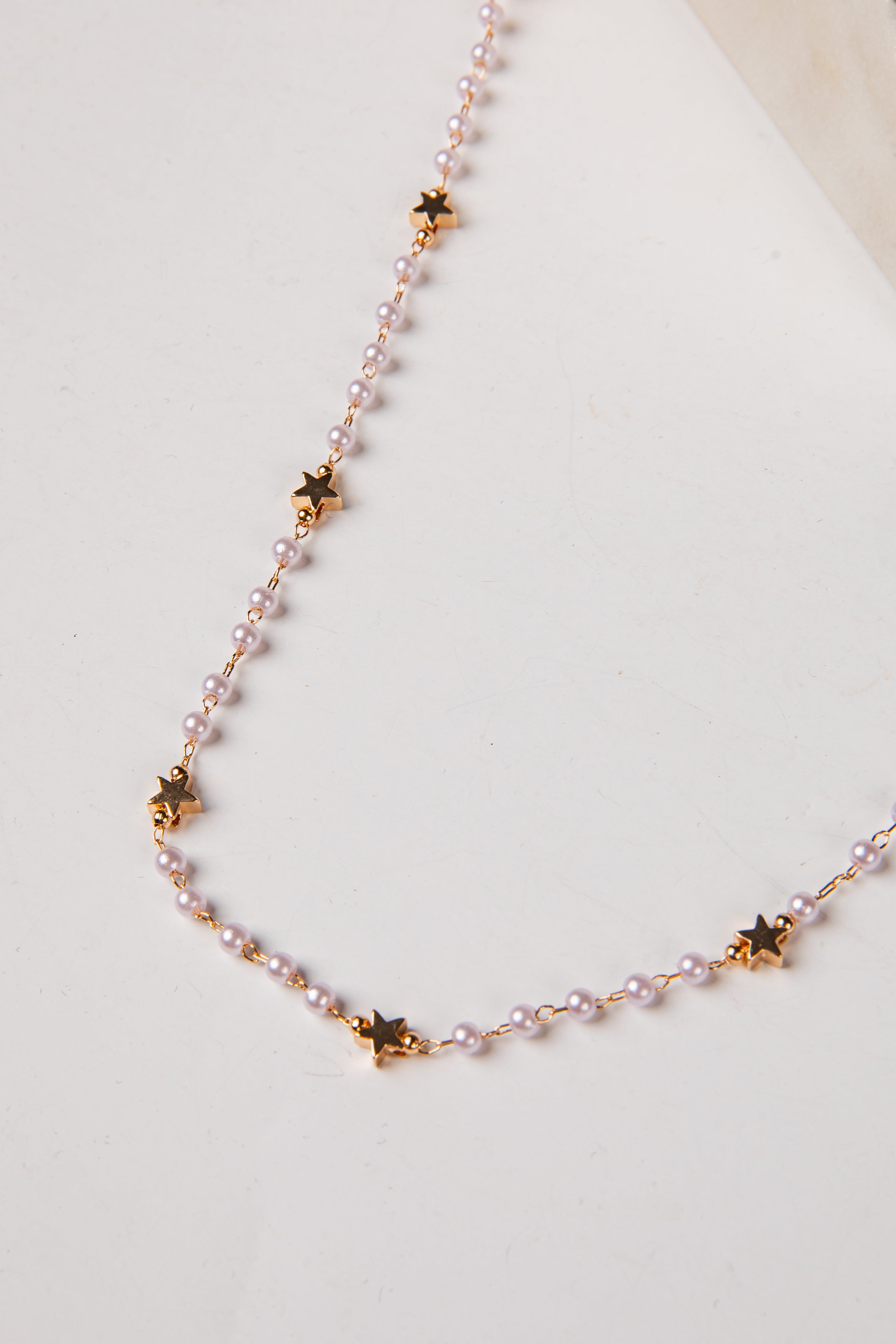 A delicate necklace featuring small light pink pearls and gold star-shaped charms, displayed on a clean off-white surface.