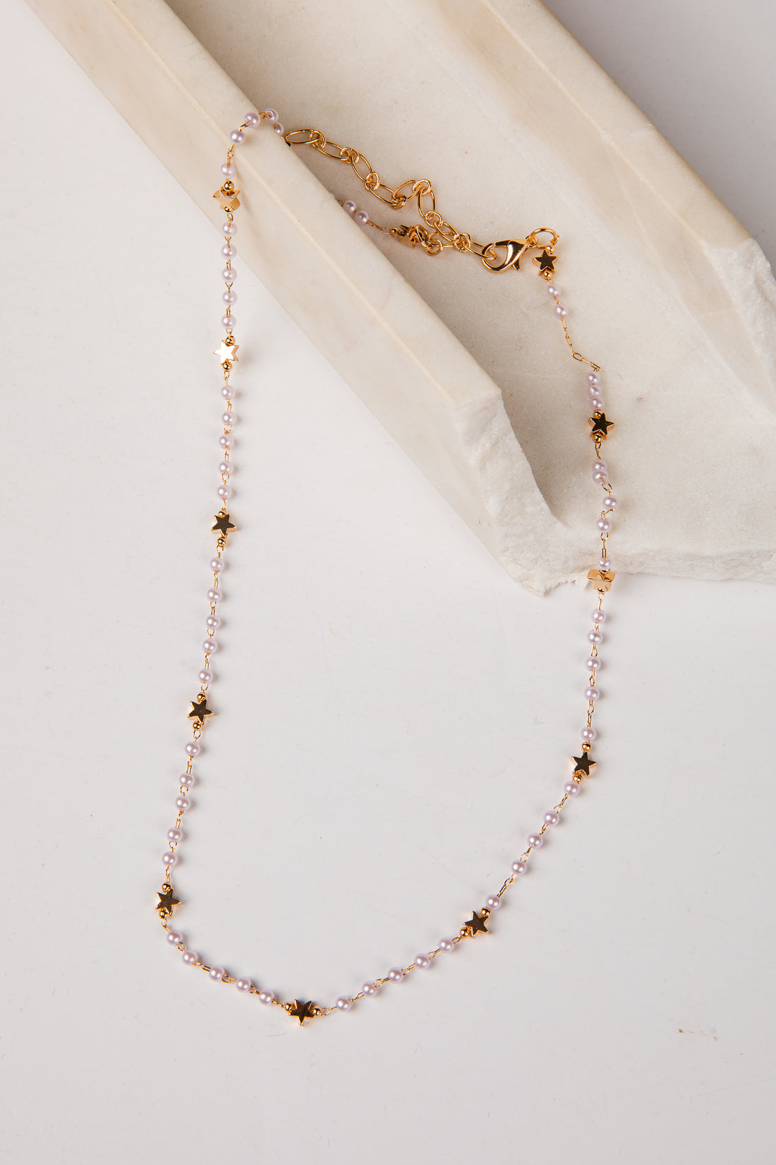 A delicate necklace with small pale pink beads and gold star-shaped accents, draped over a cream-colored fabric prop on a light background.
