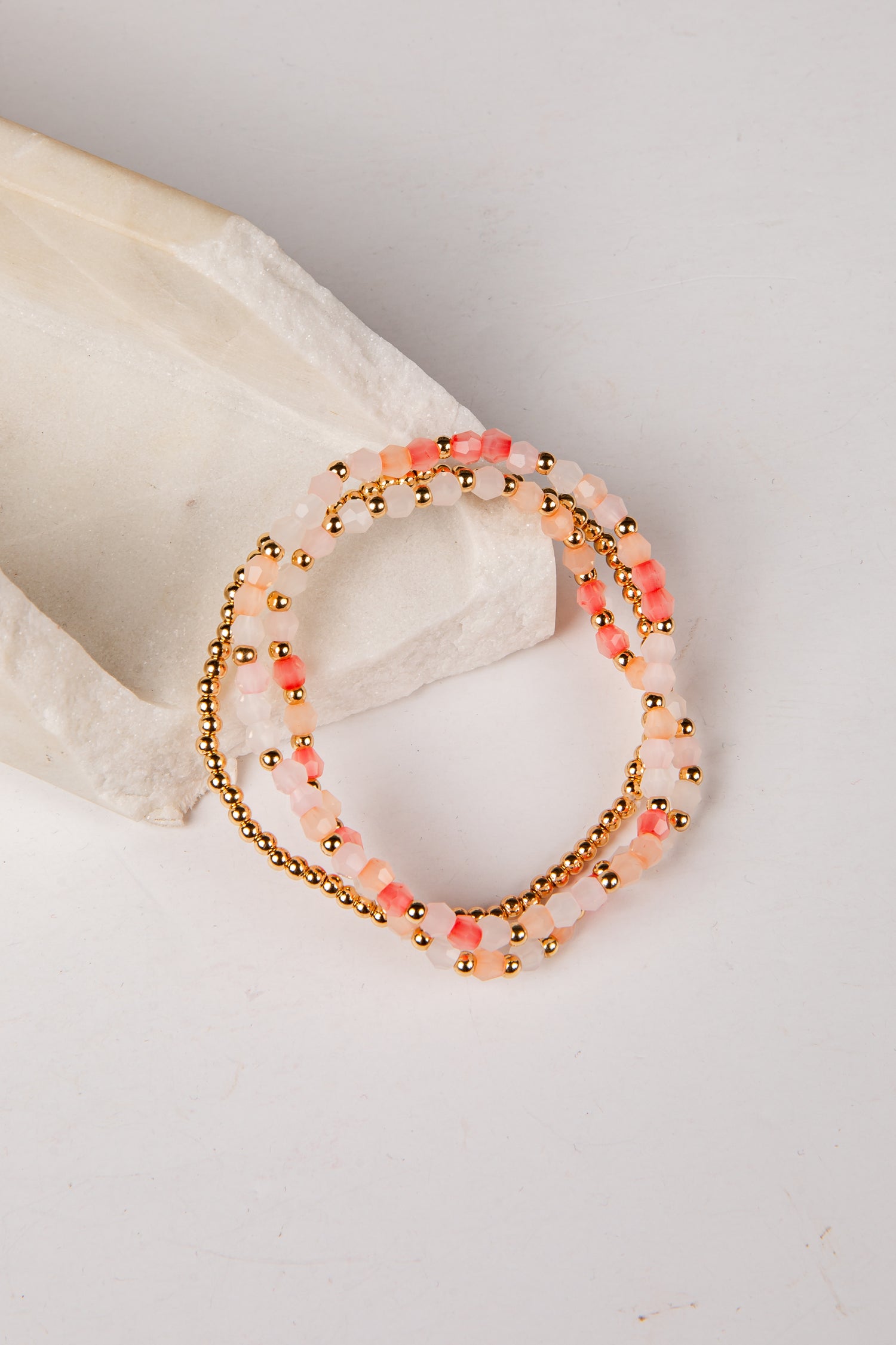 A set of three beaded stretch bracelets with pink and white beads, gold metallic beads, and coral accents, displayed on a white textured stone prop against a white background.