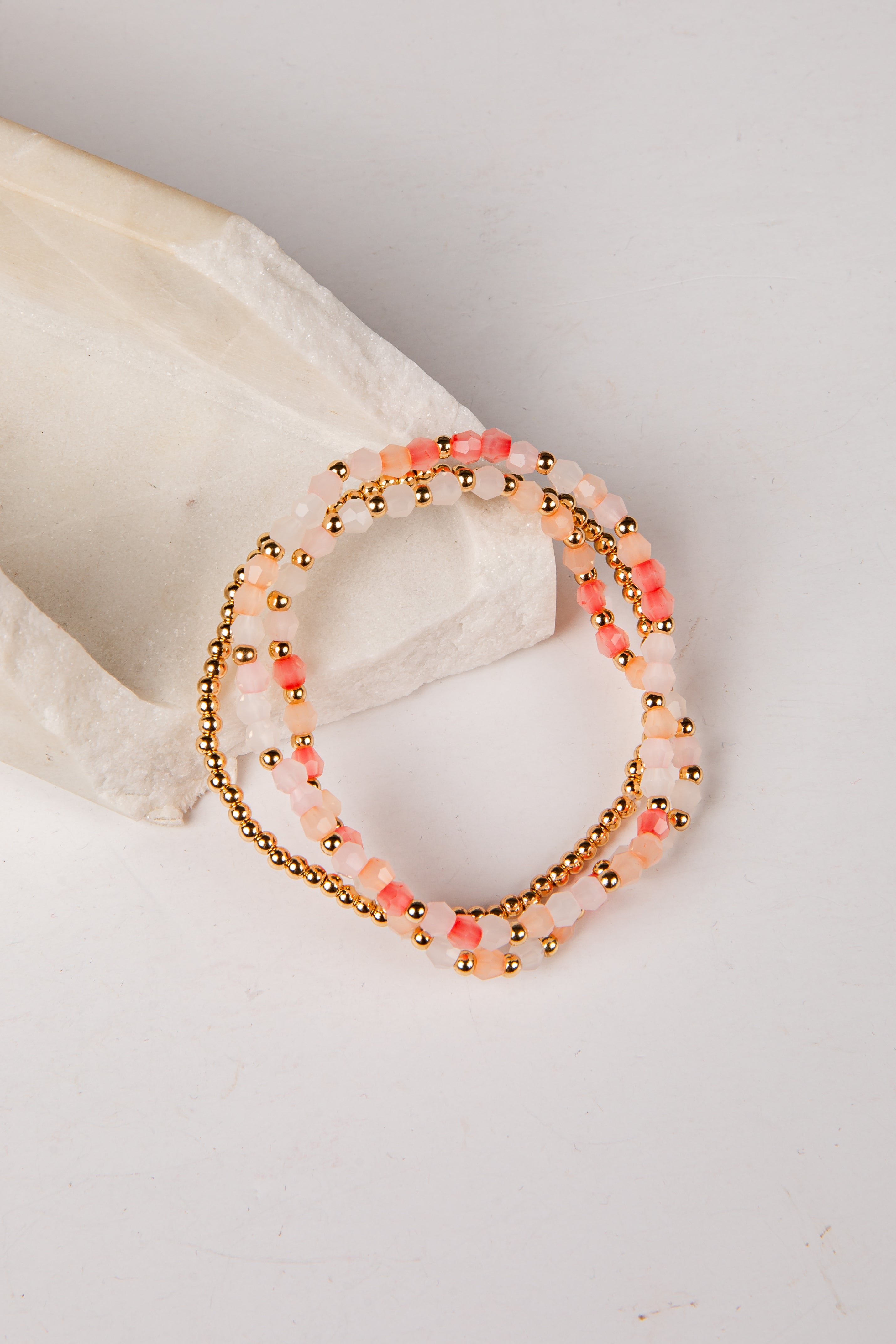 A set of three beaded stretch bracelets with pink and white beads, gold metallic beads, and coral accents, displayed on a white textured stone prop against a white background.