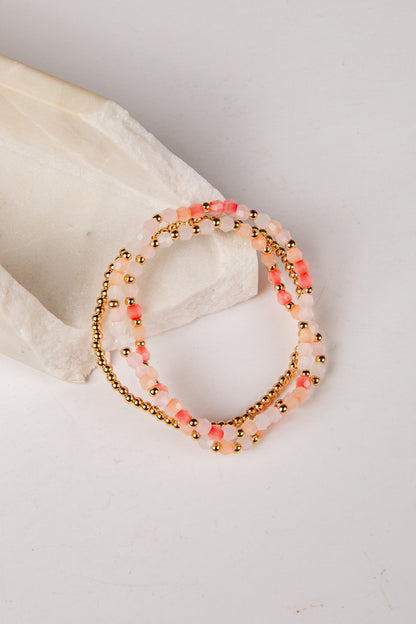 A set of three beaded stretch bracelets with pink and white beads, gold metallic beads, and coral accents, displayed on a white textured stone prop against a white background.