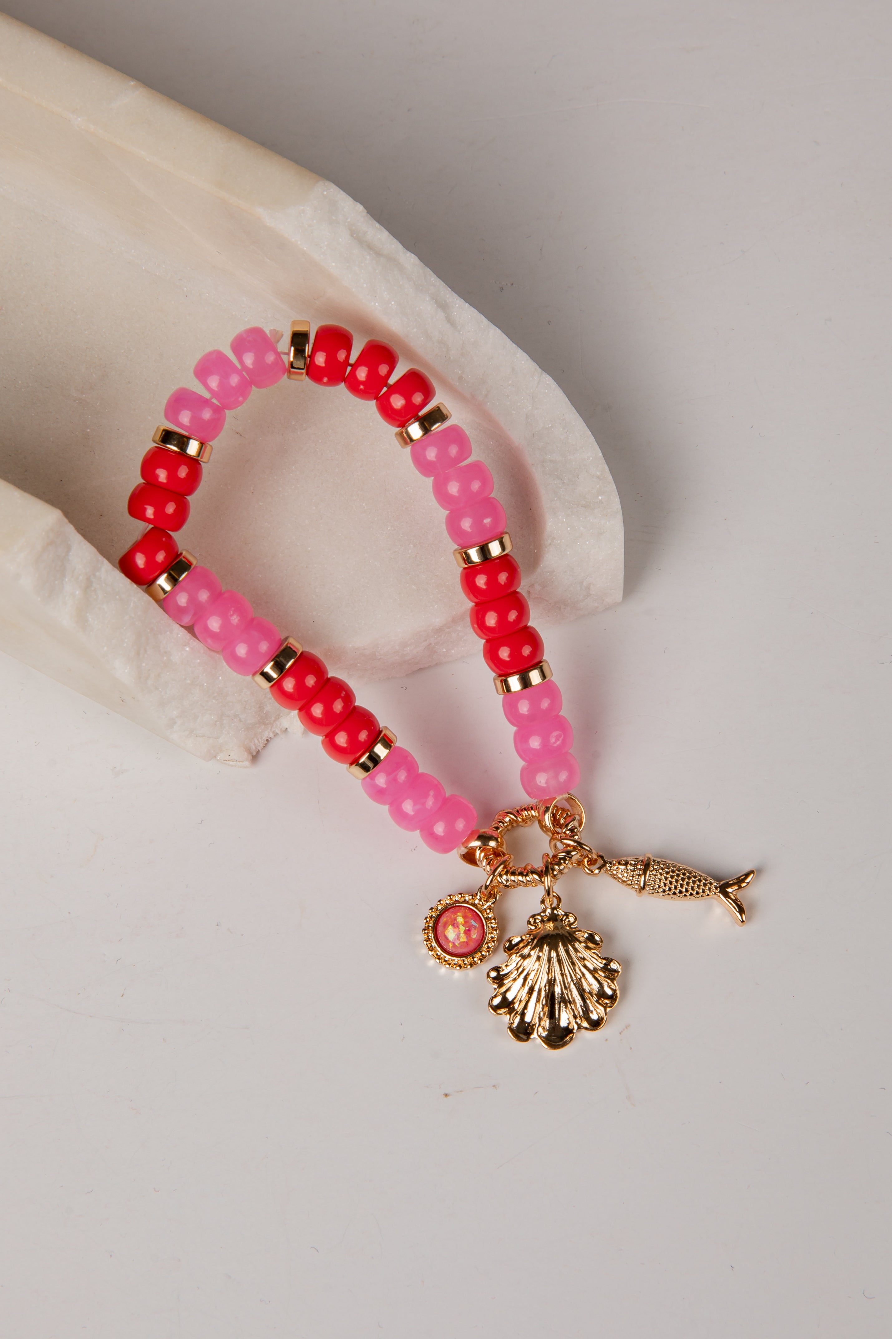 A beaded bracelet with alternating pink and red beads, accented by small gold spacer beads and a dangling gold-toned shell and fish charm, displayed on a light neutral surface.