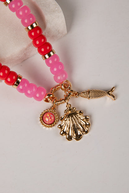 A close-up of a beaded bracelet featuring translucent pink and red beads with gold spacer rings, adorned with three dangling gold charms: a seashell, a fish, and a round medallion.