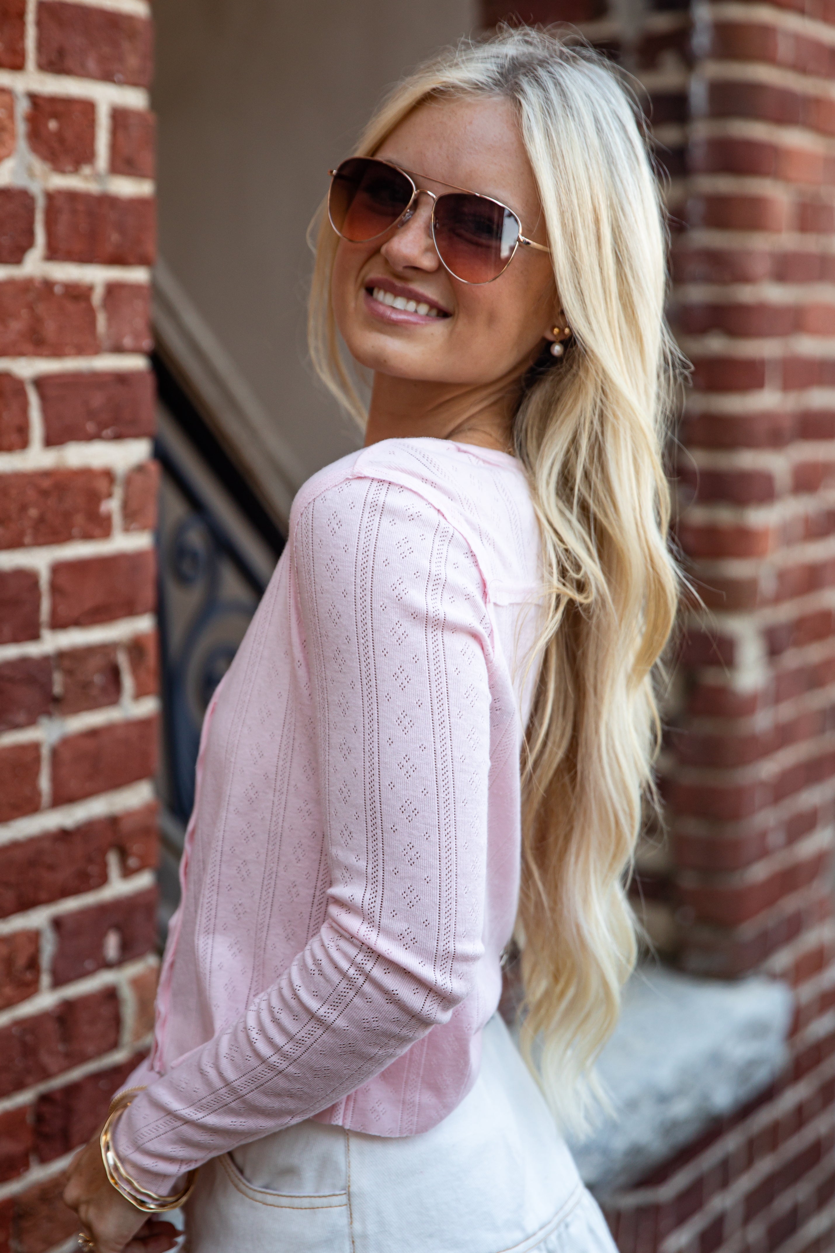 A blonde woman wearing a light pink ribbed long-sleeve top and white jeans, looking back over her shoulder while wearing aviator sunglasses in front of a red brick wall.