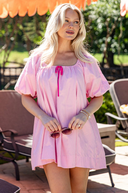 Woman in a pink dress standing outdoors with greenery and furniture in the background