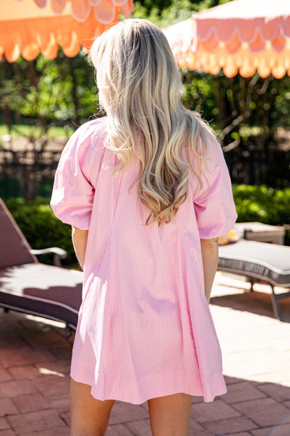 Woman in a pink dress standing outdoors with chairs and an umbrella in the background