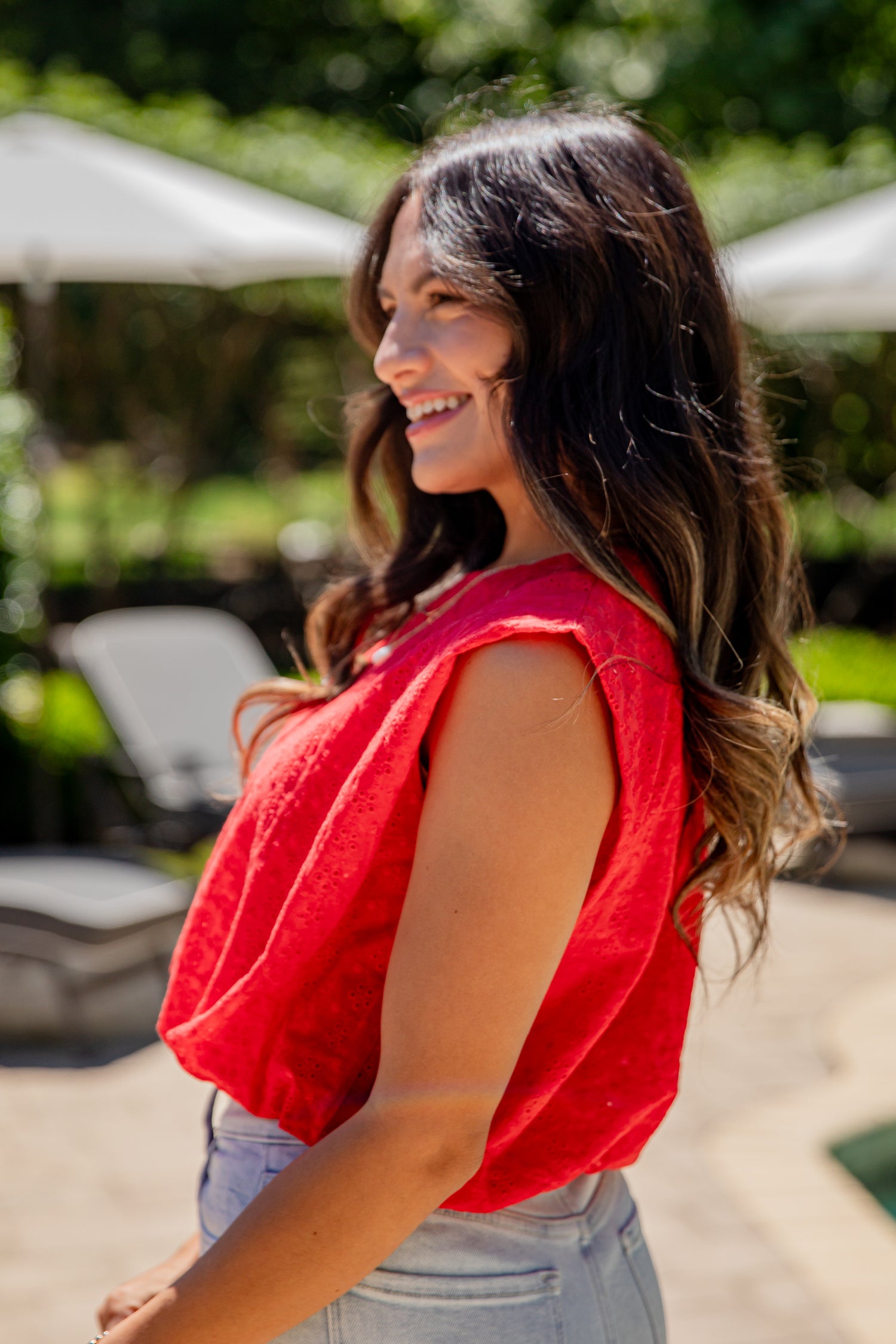 Woman in a red top standing outdoors with greenery and lounge chairs in the background