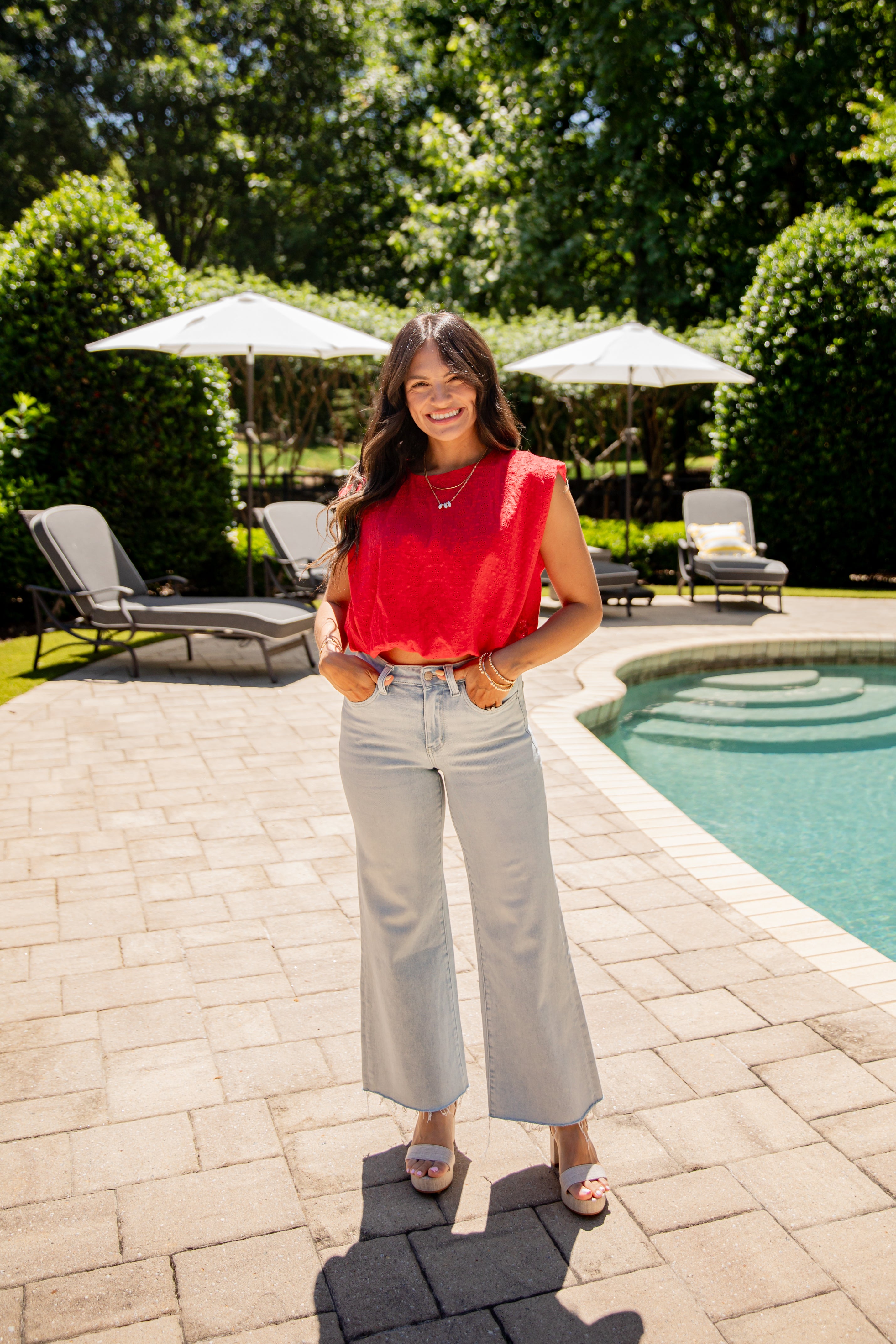 Woman in a red top and light blue jeans standing by a pool with greenery in the background
