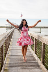 Woman in a red and white checkered dress standing on a wooden dock by a lake.