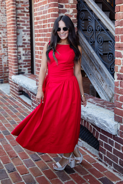 A woman wearing a sleeveless bright red midi dress and white heeled sandals stands on a brick walkway in front of a brick building with a staircase.