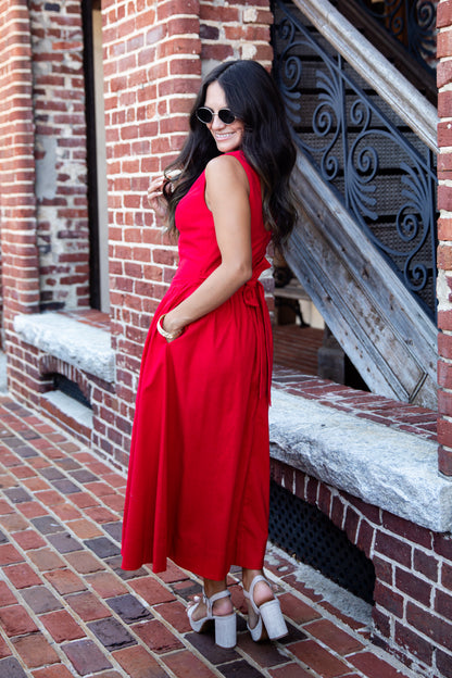 A woman wearing a sleeveless red midi dress and white platform sandals stands on a brick sidewalk beside a red brick building with a staircase.