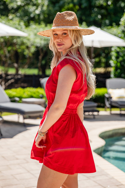 Woman in a red dress and straw hat standing by a poolside.