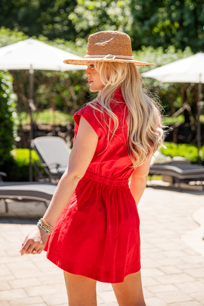 Woman in a red dress and straw hat standing by a poolside.