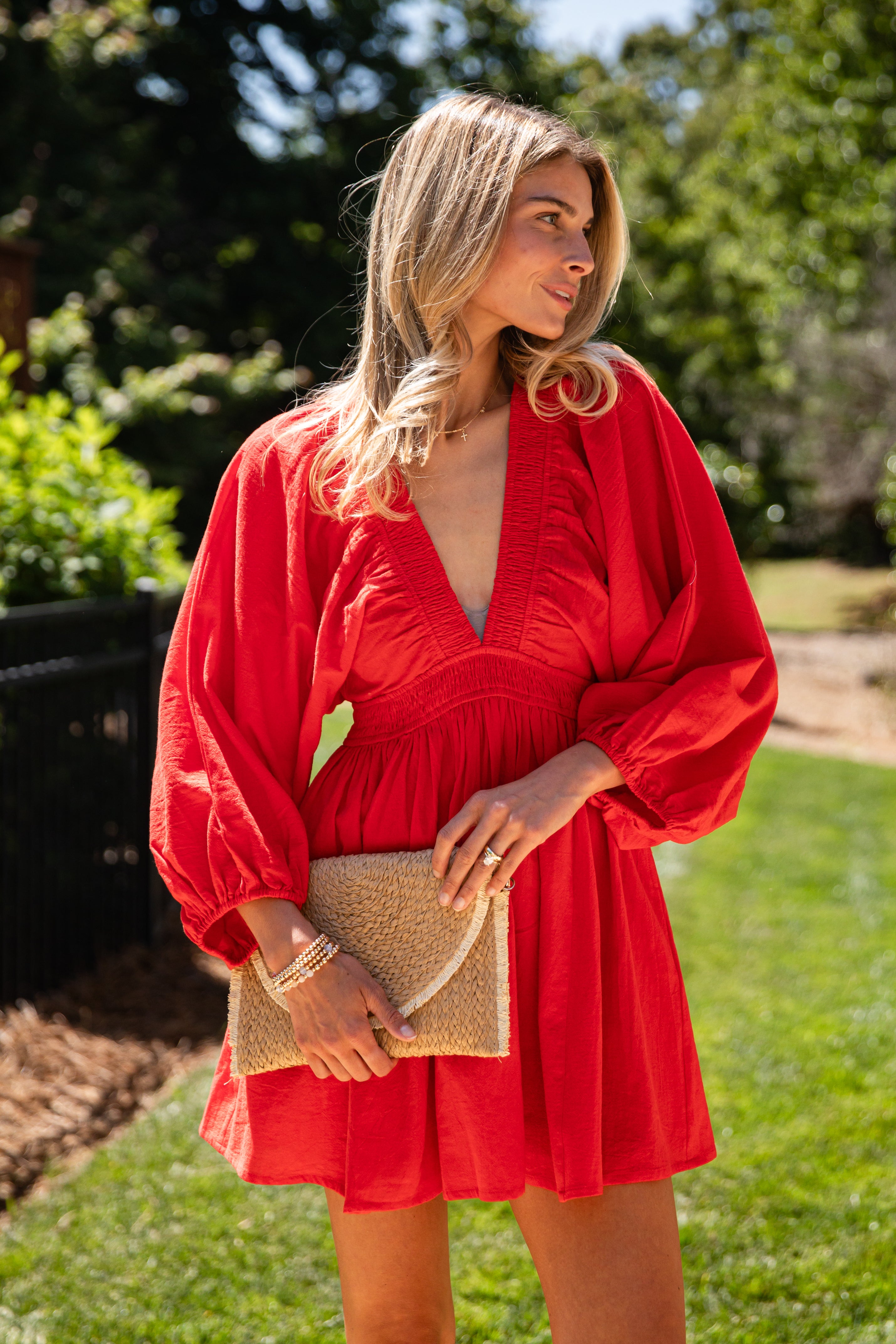 A woman wearing a bright red long-sleeve mini dress with a deep V-neck and gathered waist, holding a woven beige clutch outdoors on a grassy lawn with trees in the background.