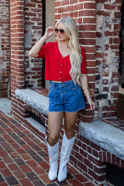 A woman stands outdoors against a red brick building, wearing a bright red short-sleeve button-front top, blue denim shorts, and white cowboy boots.