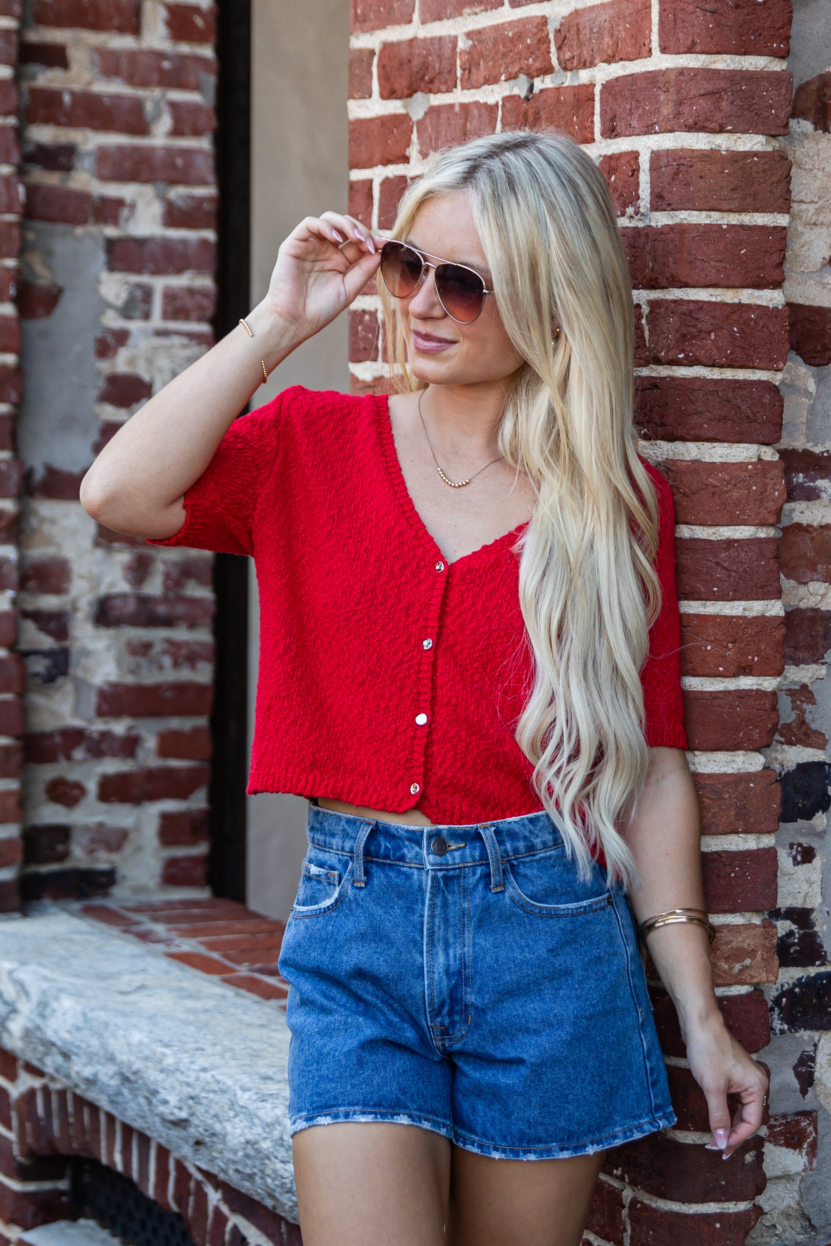 A blonde woman wearing a red short-sleeve knit cardigan and blue denim shorts, posing against a red brick wall.