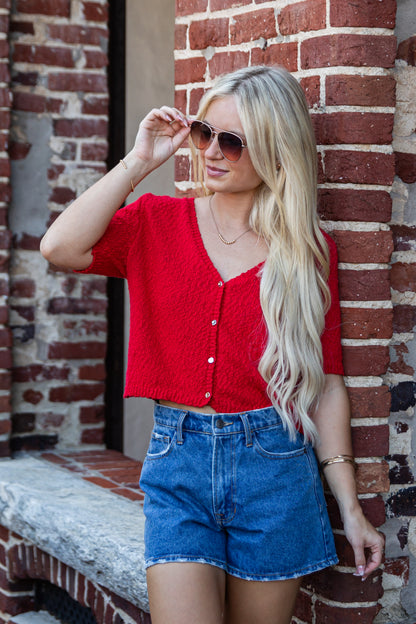 A blonde woman wearing a red short-sleeve knit cardigan and blue denim shorts, posing against a red brick wall.